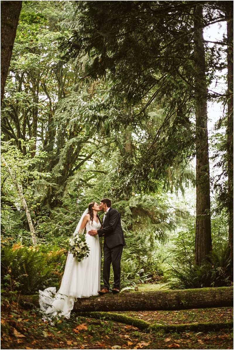 A bride and groom kiss while standing on a fallen tree trunk in a lush green snohomish forest The bride wears a white dress and holds a bouquet the groom wears a dark suit Tall trees and greenery surround them Snohomish wedding photography
