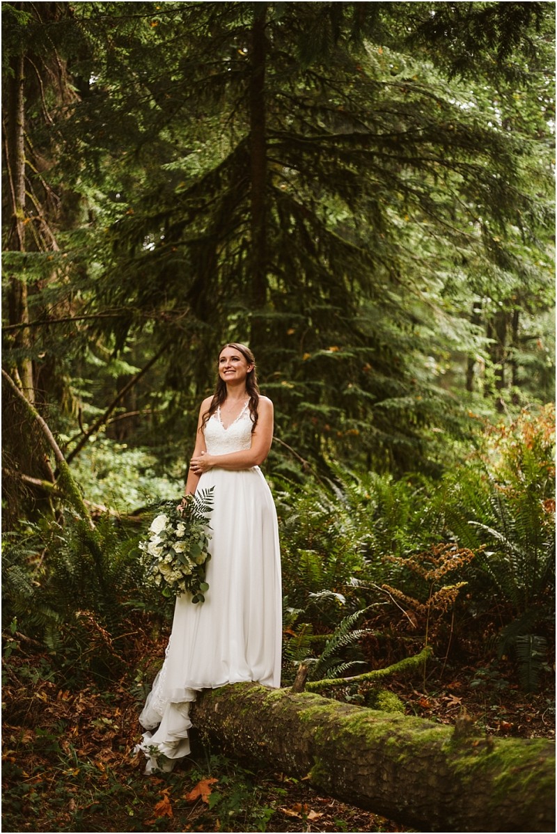 A bride in a white dress stands on a mossy log in a lush snohomish forest holding a bouquet of white flowers and greenery surrounded by tall trees and ferns Snohomish wedding photography