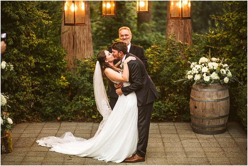 A bride and groom share a kiss during their outdoor snohomish wedding ceremony standing before an officiant They are surrounded by greenery lanterns and floral arrangements with a wine barrel and white flowers nearby Snohomish wedding photography