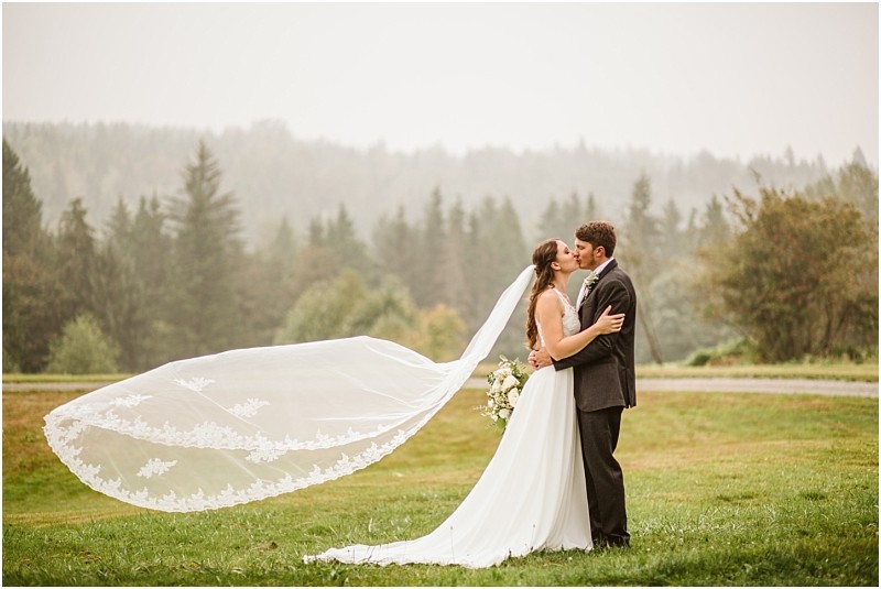 A bride and groom kiss outdoors on a grassy snohomish field surrounded by trees The brides long lace veil flows dramatically in the wind as she holds a bouquet and the groom embraces her Snohomish wedding photography