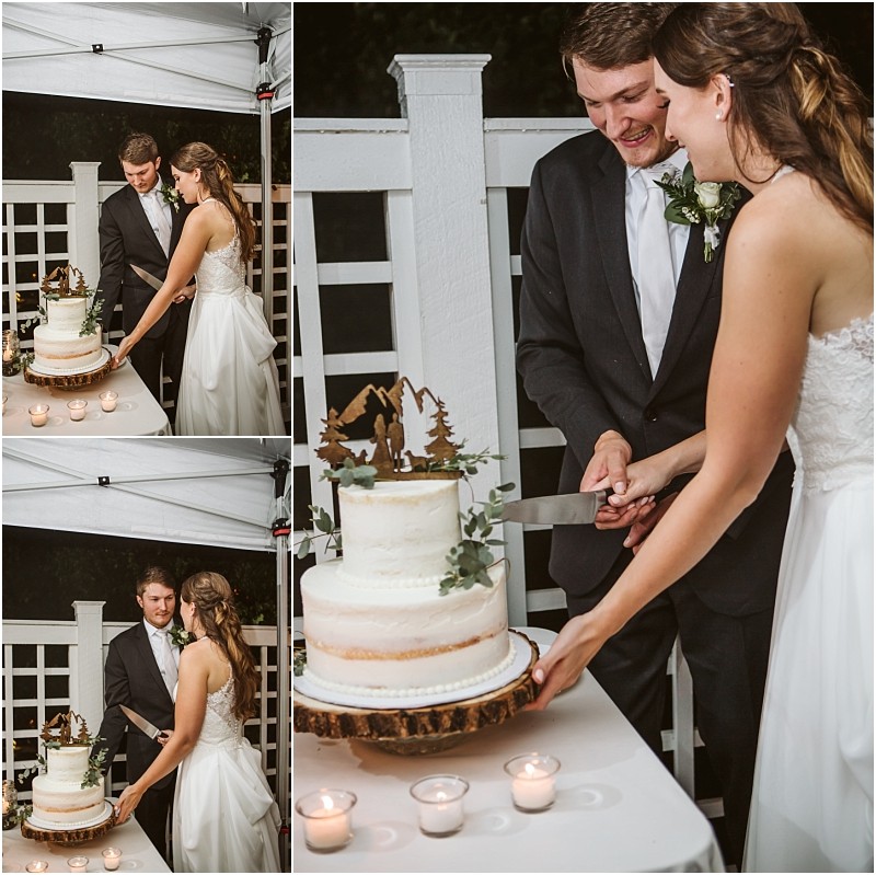 A bride and groom dressed in wedding attire smile as they cut a white rustic style wedding cake together outdoors in snohomish The cake is decorated with greenery and a nature themed topper Three candles sit on the table Snohomish wedding photography
