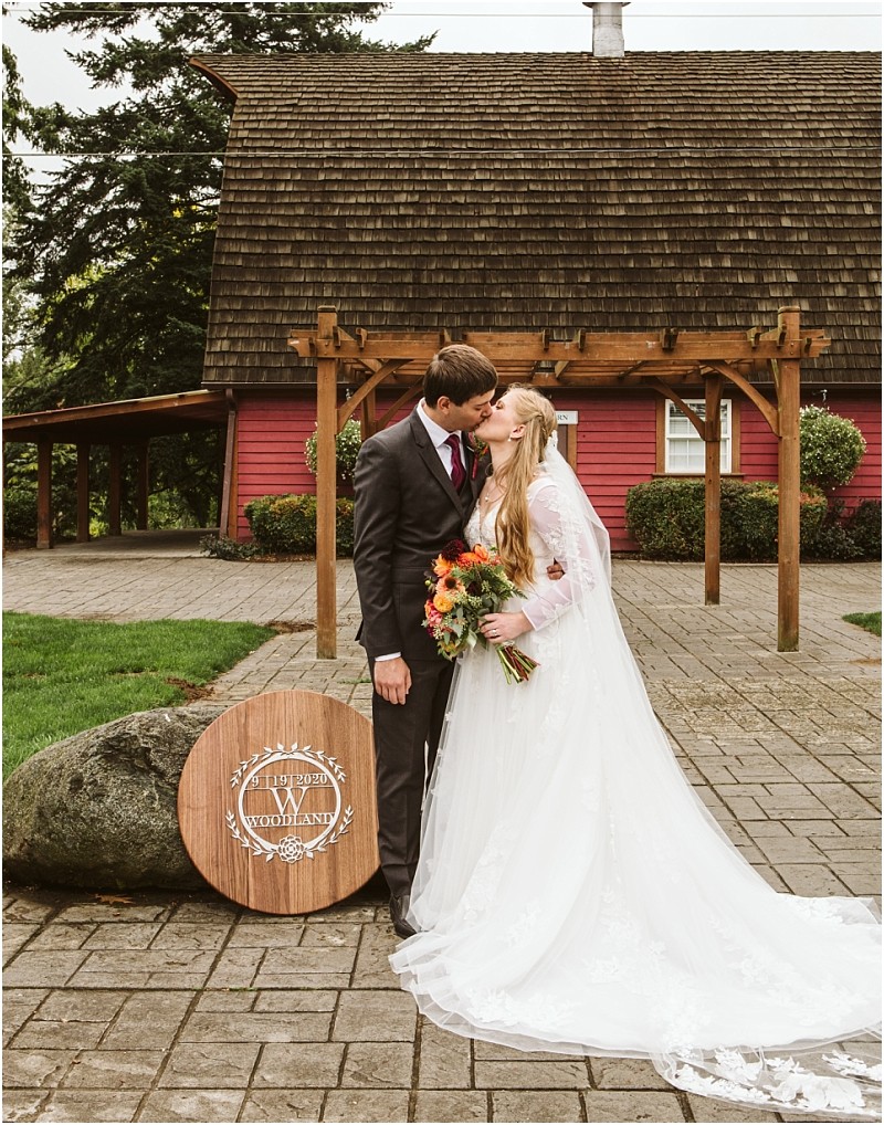 A bride and groom share a kiss outside a rustic red barn in snohomish The bride wears a white gown and holds a bouquet while the groom is in a dark suit A round wooden sign reads the estates at woodland in front of them Snohomish wedding photography
