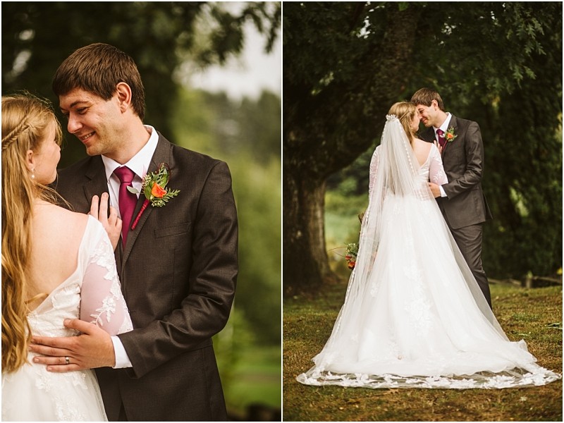 A bride and groom stand outdoors under snohomish trees The groom wears a dark suit with a red tie and the bride wears a long sleeved lace gown and veil They embrace and smile at each other holding a small bouquet Snohomish wedding photography