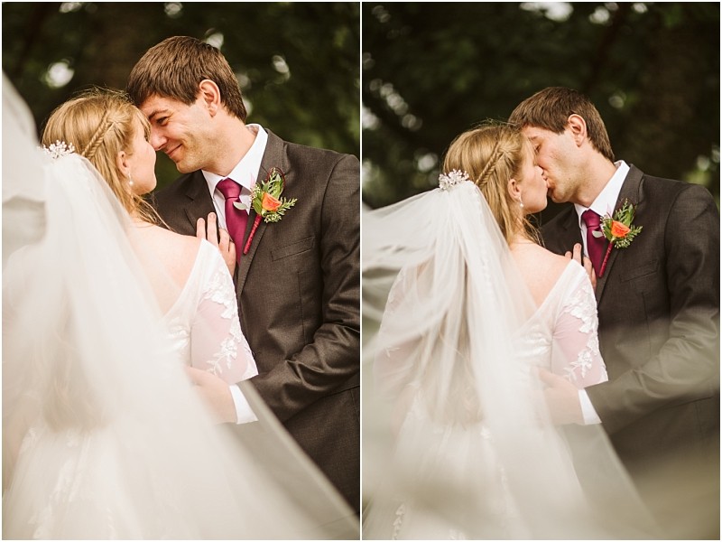 A bride and groom stand close together outdoors in snohomish The groom wears a dark suit and red tie while the bride wears a white dress with lace sleeves and a long flowing veil They share an intimate moment smiling and kissing Snohomish wedding photography