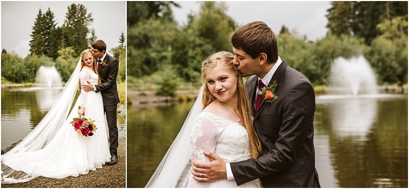 A bride and groom embrace beside a peaceful snohomish lake with fountains and trees in the background The groom kisses the brides head as she smiles holding a bouquet of red flowers and wearing a long white wedding dress and veil Snohomish wedding photography
