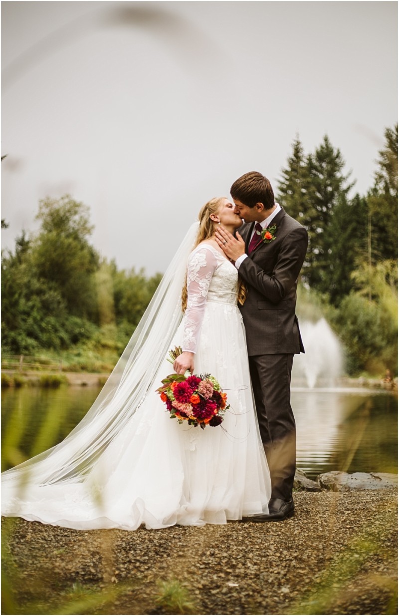 A bride in a white gown and a groom in a dark suit share a kiss by a snohomish lakeside with trees and a fountain in the background The bride holds a bouquet of red flowers and wears a long veil Snohomish wedding photography