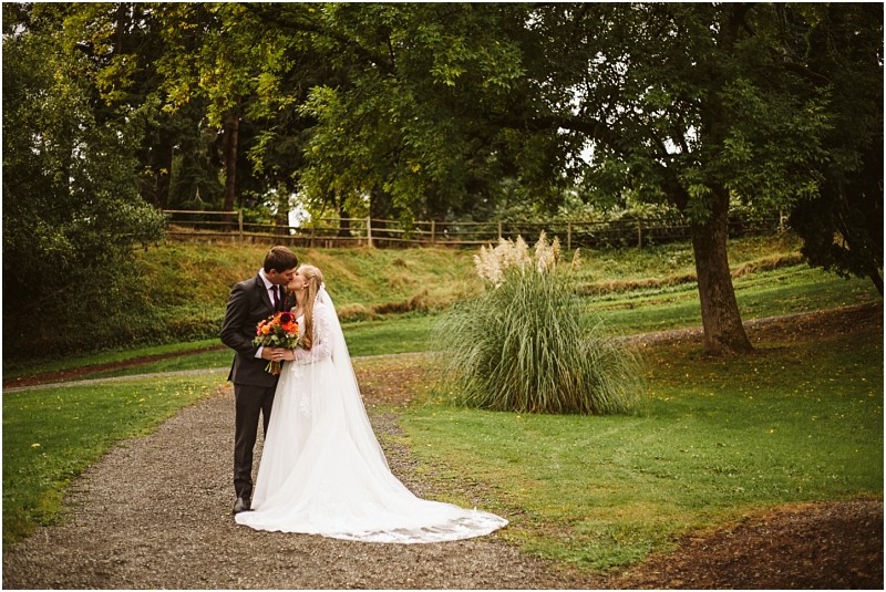 A bride and groom share a kiss on a gravel path surrounded by snohomish greenery The bride wears a long white dress and veil holding a bouquet of flowers amid the peaceful outdoor setting of trees and grass Snohomish wedding photography
