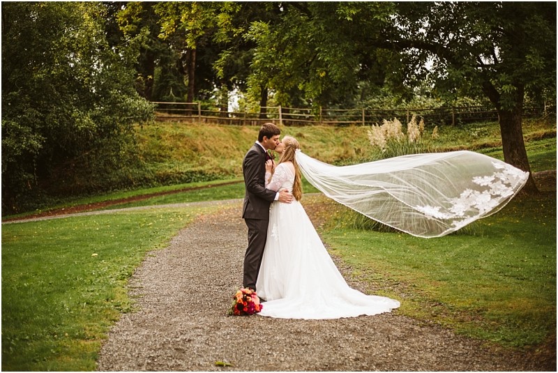 A bride and groom stand on a gravel path in a snohomish park embracing and kissing The brides long veil flows in the wind and a bouquet of flowers rests at their feet amid lush trees and greenery Snohomish wedding photography