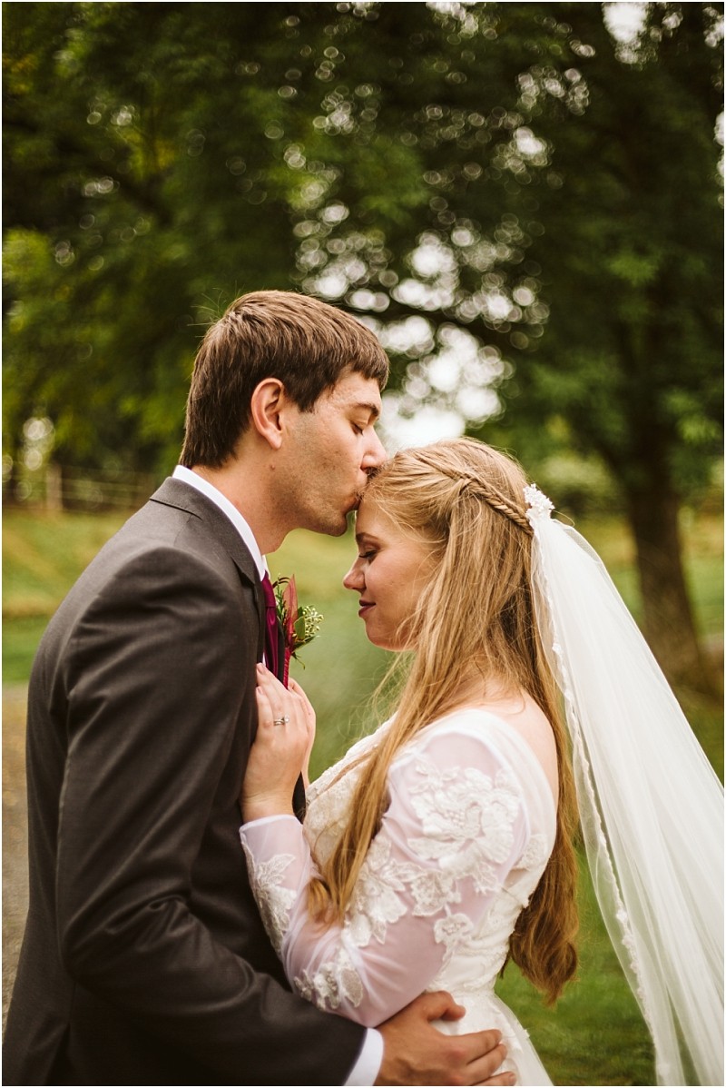 A groom in a dark suit kisses the forehead of his bride who wears a white lace dress and veil as they embrace outdoors with green trees in the background of snohomish Snohomish wedding photography