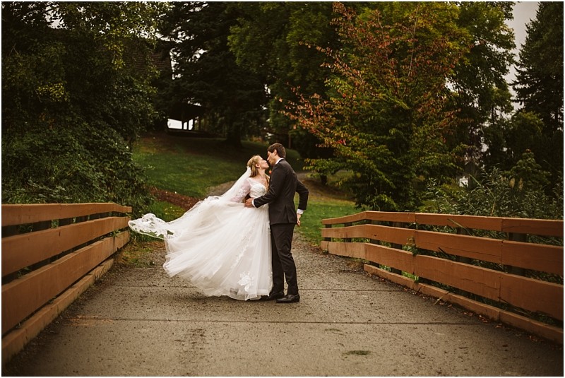 A bride and groom share a kiss on a wooden bridge in snohomish surrounded by lush trees and greenery The brides white dress flows behind her as the couple stands together on a gray overcast day Snohomish wedding photography