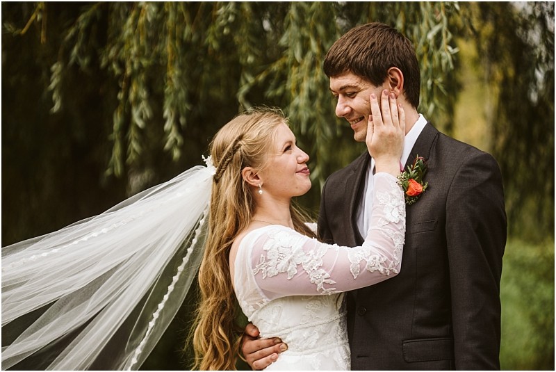A bride in a white lace dress and veil smiles lovingly as she touches the face of the groom who is wearing a dark suit with an orange boutonniere They stand close together outdoors amid lush snohomish greenery Snohomish wedding photography