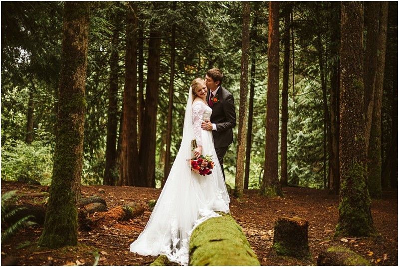 A bride in a white gown and a groom in a suit stand together in a lush green snohomish forest The groom kisses the brides head as she holds a bouquet of red flowers both standing on a mossy fallen tree Snohomish wedding photography