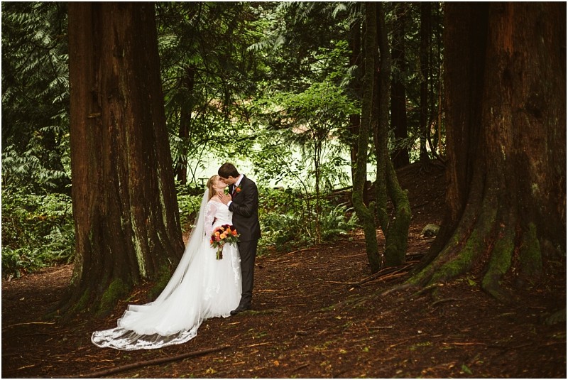 A bride and groom share a kiss in a snohomish forest surrounded by tall trees The bride wears a white dress with a long train and holds a bouquet while the groom is in a suit Natural light filters through the greenery Snohomish wedding photography