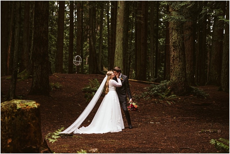 A bride and groom share a kiss in a snohomish forest surrounded by tall trees The bride wears a long white dress and veil while the groom holds a bouquet of flowers The scene is peaceful and intimate Snohomish wedding photography