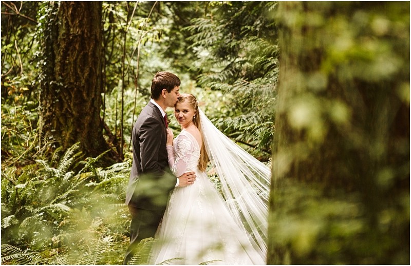 A bride and groom stand close together in a lush green snohomish forest The bride wears a white dress with a long veil and the groom is in a dark suit Sunlight filters through the trees creating a romantic serene atmosphere Snohomish wedding photography