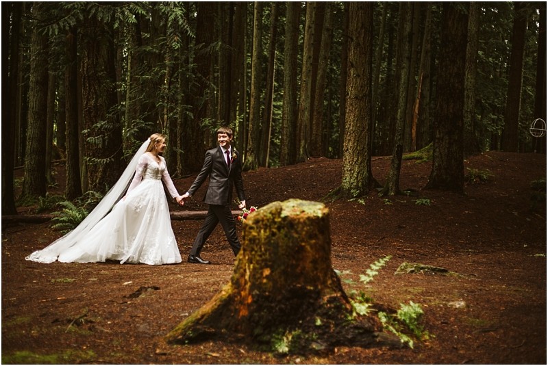 A bride and groom hold hands and walk through a snohomish forest surrounded by tall trees and mossy ground The bride wears a white wedding dress and veil while the groom wears a dark suit Snohomish wedding photography