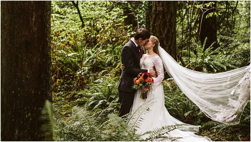 A bride and groom embrace and share a kiss in a lush green snohomish forest The bride holds a bouquet of flowers and her long veil flows behind her creating a romantic scene among the trees and ferns Snohomish wedding photography