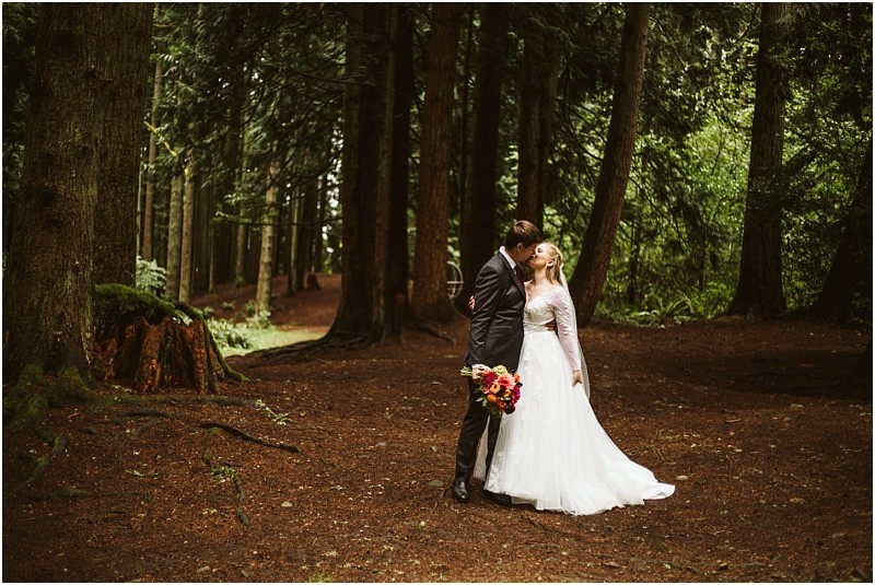 A bride and groom stand kissing in a snohomish forest surrounded by tall trees The bride wears a long white dress and holds a bouquet of flowers while the groom is in a dark suit Soft light filters through the trees Snohomish wedding photography