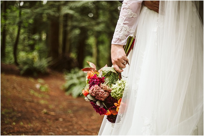 A bride in a white lace wedding dress holds a colorful bouquet of flowers in a snohomish forest setting The focus is on her hand and the bouquet with greenery and trees softly blurred in the background Snohomish wedding photography