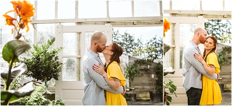 A couple embraces and kisses inside a bright snohomish greenhouse The man in a light blue shirt holds a woman in a yellow dress as sunlight streams through the glass windows with green plants and orange flowers in the foreground Snohomish wedding photography