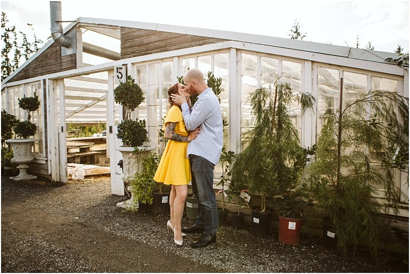 A couple shares a kiss in front of a snohomish greenhouse The woman wears a bright yellow dress and heels while the man wears a light blue shirt and jeans Potted plants line the entrance to the rustic glass building Snohomish wedding photography