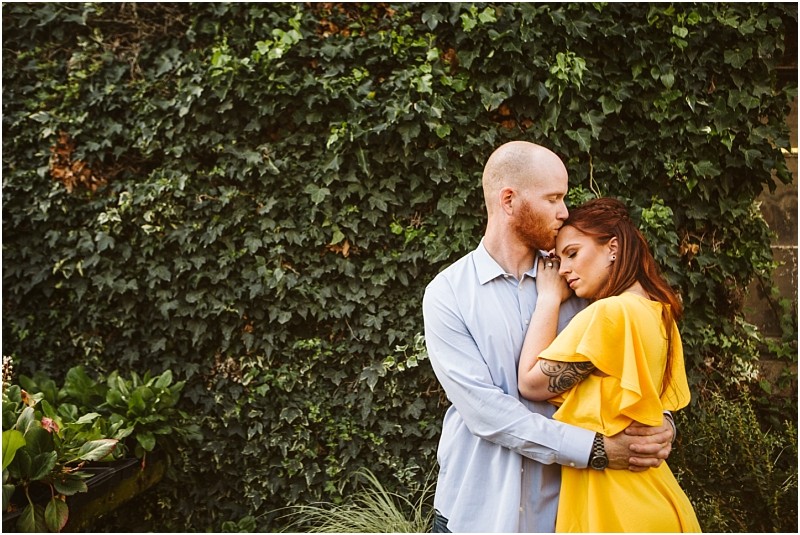 A man embraces a woman in a yellow dress gently kissing her forehead as she rests her head on his chest They stand in front of a lush ivy covered wall surrounded by greenery in picturesque snohomish Snohomish wedding photography
