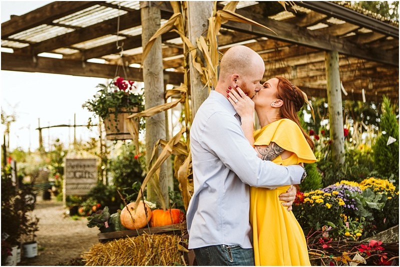 A couple embraces and kisses in a rustic snohomish outdoor setting surrounded by flowers pumpkins and cornstalks The woman wears a yellow dress and the man wears a light blue shirt The scene has a warm autumn feel Snohomish wedding photography