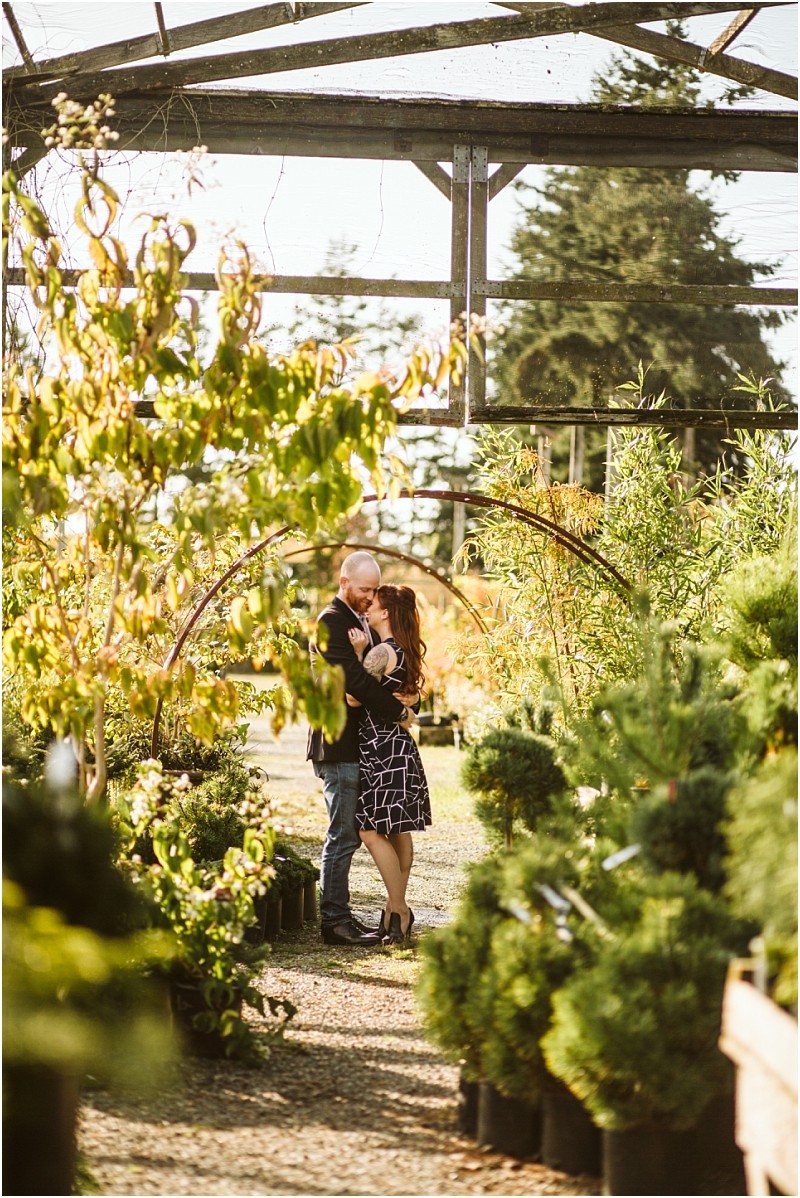 A couple stands close together embracing and smiling at each other in the middle of a sunlit snohomish plant nursery surrounded by lush green shrubs and trees The scene radiates warmth and romance Snohomish wedding photography