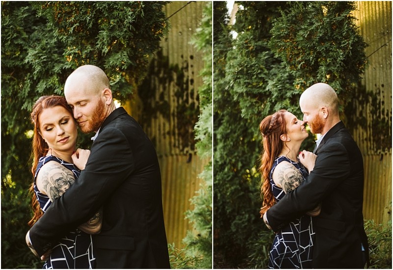 A couple stands close together outdoors in snohomish embracing and smiling at each other in front of green trees and a corrugated metal wall The woman has long red hair and tattoos the man is bald with a beard and wearing a black suit Snohomish wedding photography