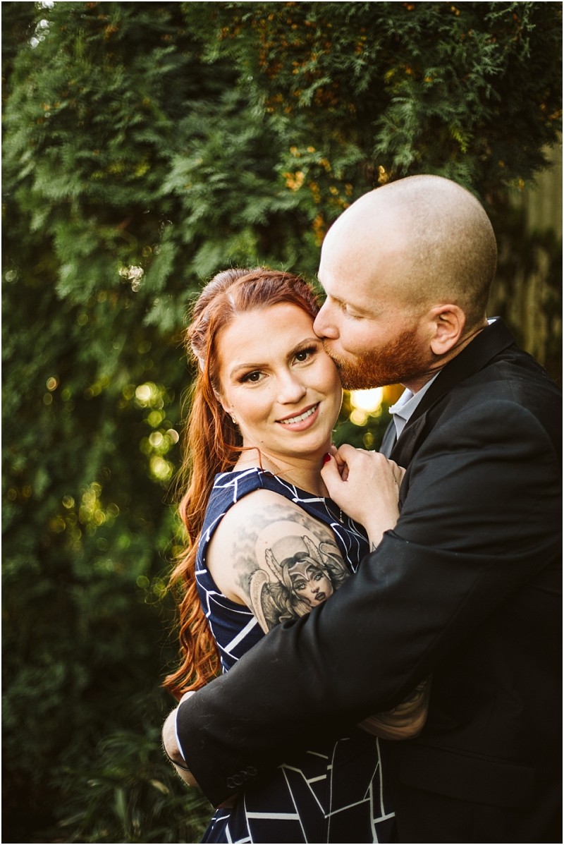 A man in a black suit gently kisses a smiling woman with long red hair and a large tattoo on her upper arm as they embrace outdoors surrounded by lush snohomish greenery Snohomish wedding photography