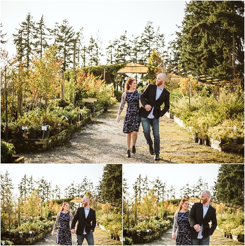 A couple walks hand in hand through a sunny snohomish plant nursery surrounded by greenery and potted plants They smile at each other dressed smartly in semi formal attire with trees and a pergola in the background Snohomish wedding photography