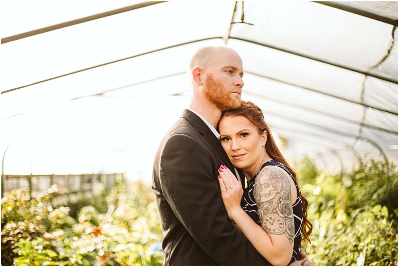A couple stands under a greenhouse structure in snohomish with the bearded man in a suit embracing a woman with long hair and a tattooed arm She leans against him looking at the camera surrounded by lush greenery and sunlight Snohomish wedding photography