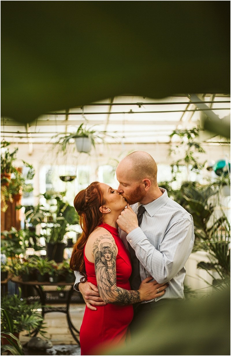 A couple shares a romantic kiss in a snohomish greenhouse surrounded by lush green plants The woman in a red dress has visible tattoos on her arm and the man dressed in a striped shirt gently holds her face Snohomish wedding photography