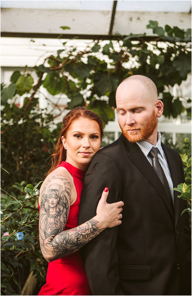 A woman in a red dress with a detailed arm tattoo embraces a man in a black suit and tie They stand close together in a snohomish greenhouse surrounded by greenery The man looks down while the woman faces the camera Snohomish wedding photography