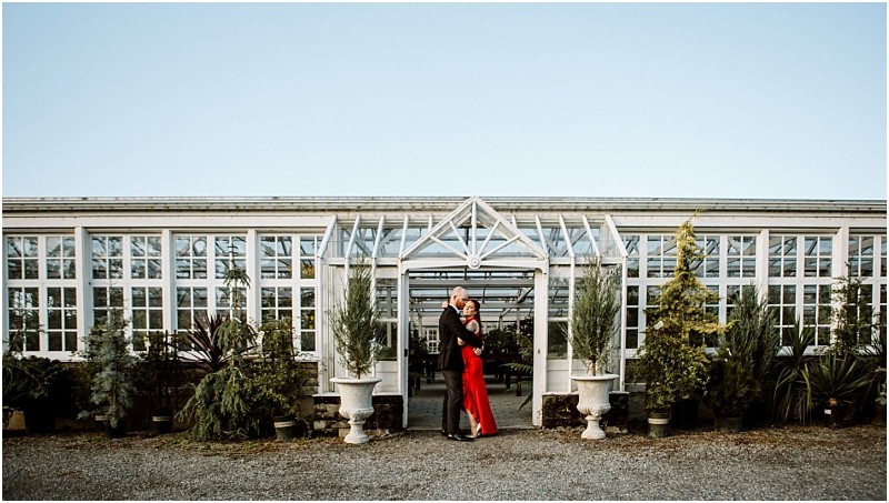 A couple embraces in front of a large glass greenhouse with white framing in snohomish surrounded by potted plants and green shrubs under a clear blue sky Snohomish wedding photography
