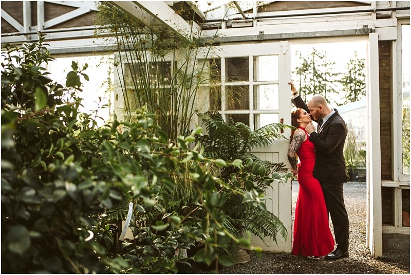 A couple dressed formally the woman in a red gown and the man in a suit share a romantic moment under a doorway in a sunlit snohomish greenhouse surrounded by lush green plants Snohomish wedding photography