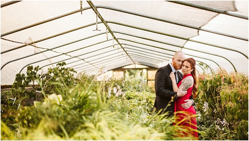 A couple embraces in a snohomish greenhouse filled with lush greenery The man wears a black suit and the tattooed woman a red dress as they stand close together surrounded by plants beneath a translucent roof Snohomish wedding photography