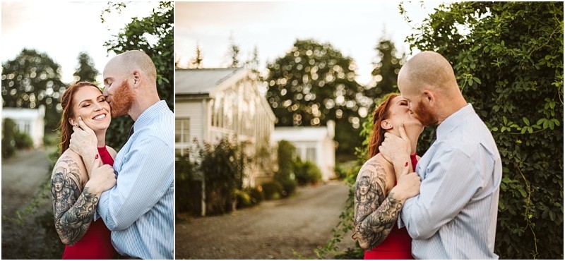 A smiling couple embraces outdoors in snohomish surrounded by greenery The woman with red hair and visible arm tattoos wears a red dress The man bald with a beard wears a light blue shirt They look into each others eyes affectionately Snohomish wedding photography