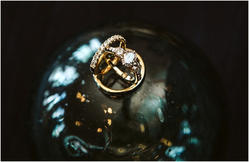 A close up of two intertwined gold rings one with a sparkling diamond resting on a reflective dark surface with soft blurred lights in the backgroundcapturing the romantic essence of snohomish Snohomish wedding photography
