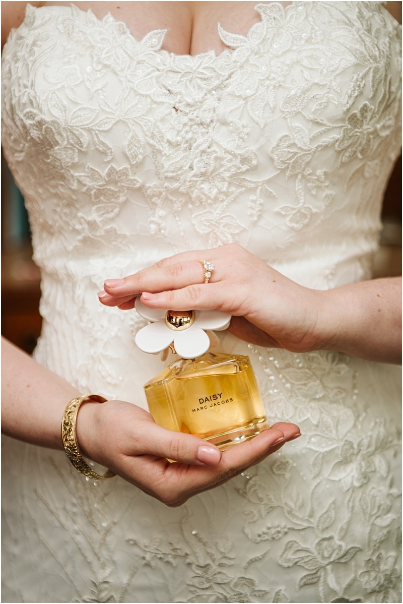 A bride in a white lace wedding dress holds a bottle of daisy by marc jacobs perfume at her snohomish wedding Her hands display a gold bracelet and engagement ring while the bottles cap is adorned with white flower shaped decorations Snohomish wedding photography