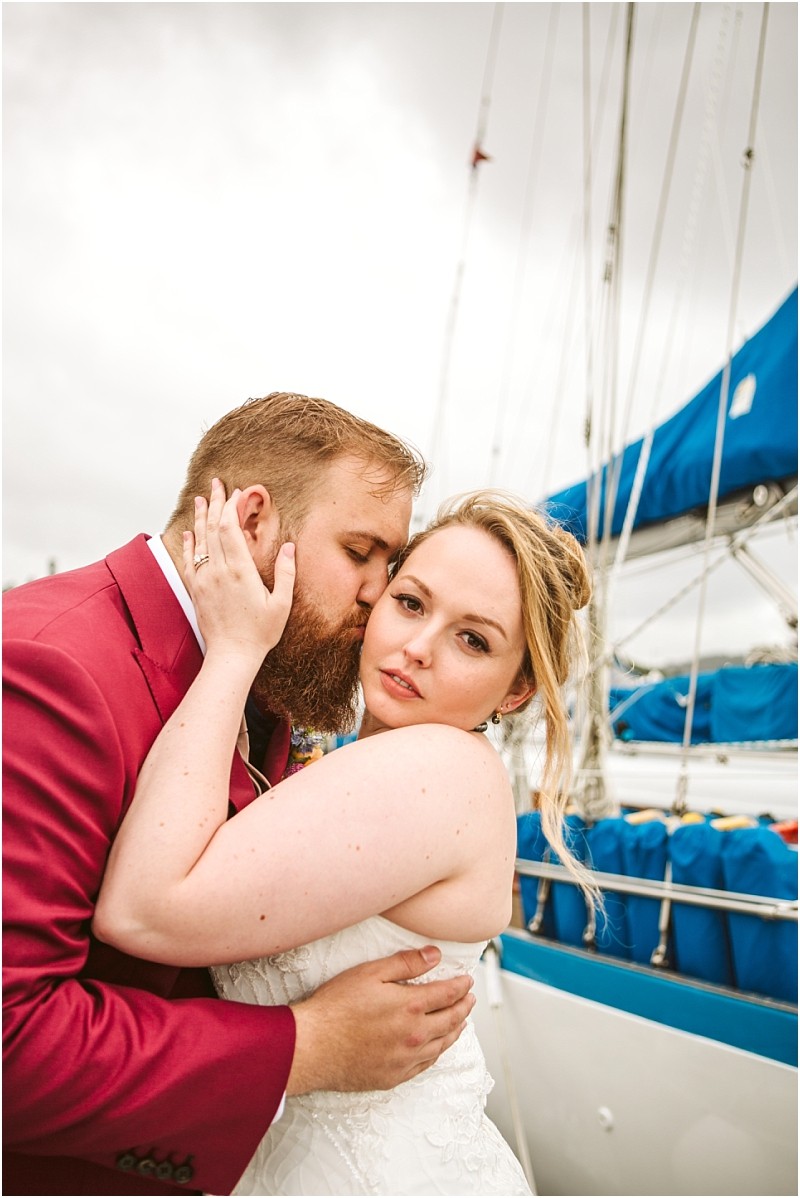 A groom in a burgundy suit kisses the cheek of a bride in a white dress as she looks at the camera standing close together on a docked sailboat with blue covers in snohomish Snohomish wedding photography