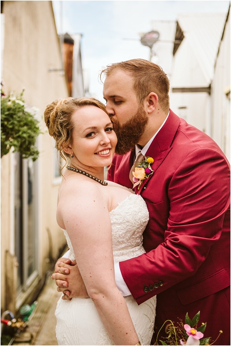 A groom in a red suit kisses his brides forehead as she smiles at the camera They embrace in a narrow snohomish alleyway surrounded by lush greenery and charming historic buildings Snohomish wedding photography