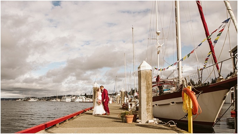 A bride and groom stand together on a dock beside sailboats under a cloudy snohomish sky with water and distant buildings visible in the background Snohomish wedding photography