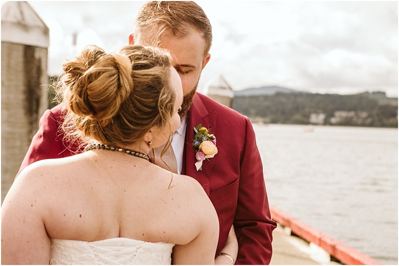 A couple stands close together on a snohomish dock by the water The woman in a strapless white dress with her hair in a bun faces the man in a maroon suit and boutonniere Their heads are touching affectionately Snohomish wedding photography