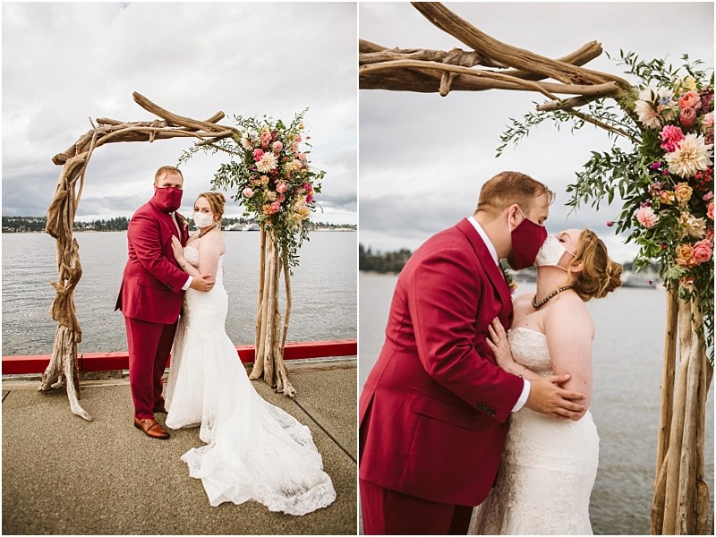 A bride and groom both wearing masks embrace and kiss under a rustic wooden wedding arch decorated with pink flowers by the snohomish waterfront on a cloudy day The groom wears a red suit the bride wears a white strapless gown Snohomish wedding photography