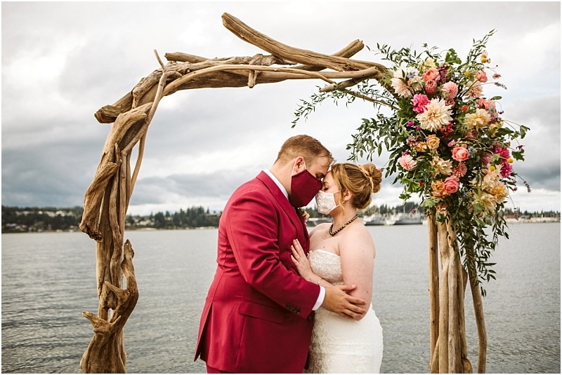 A bride and groom both wearing face masks embrace under a rustic wooden wedding arch decorated with colorful flowers in snohomish with a lake and cloudy sky in the background Snohomish wedding photography