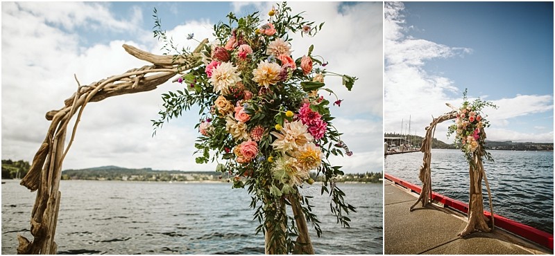 A wooden arch decorated with pink peach and white flowers stands on a dock beside a snohomish lake with boats and distant hills visible under a partly cloudy sky Snohomish wedding photography
