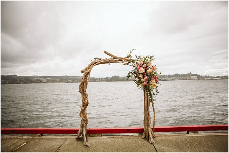 A rustic wedding arch made of driftwood decorated with pink and white flowers stands on a pier beside a large body of water under a cloudy snohomish sky Snohomish wedding photography