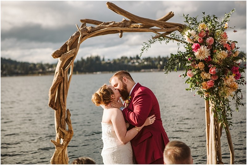 A bride and groom share a kiss under a rustic wooden arch decorated with colorful flowers with a lake and trees in the background on a cloudy snohomish day Snohomish wedding photography