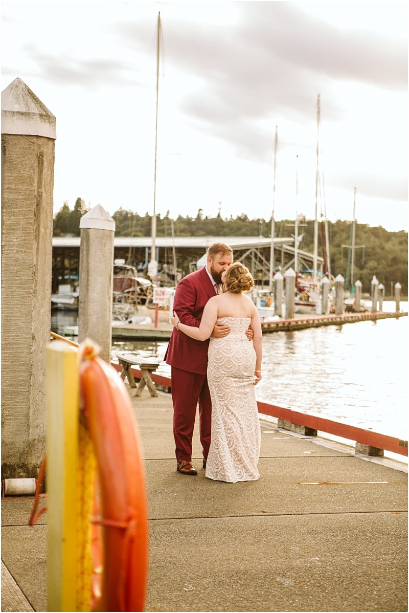A bride in a strapless lace dress and a groom in a burgundy suit embrace on a dock near the water in snohomish with sailboats and a marina in the background at sunset Snohomish wedding photography